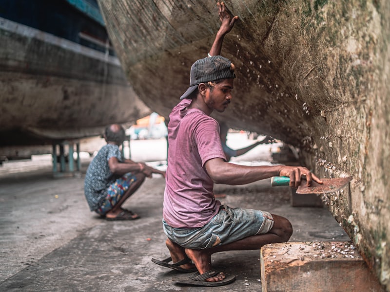 Two workers cleaning and scraping the underside of a hull in a boatyard